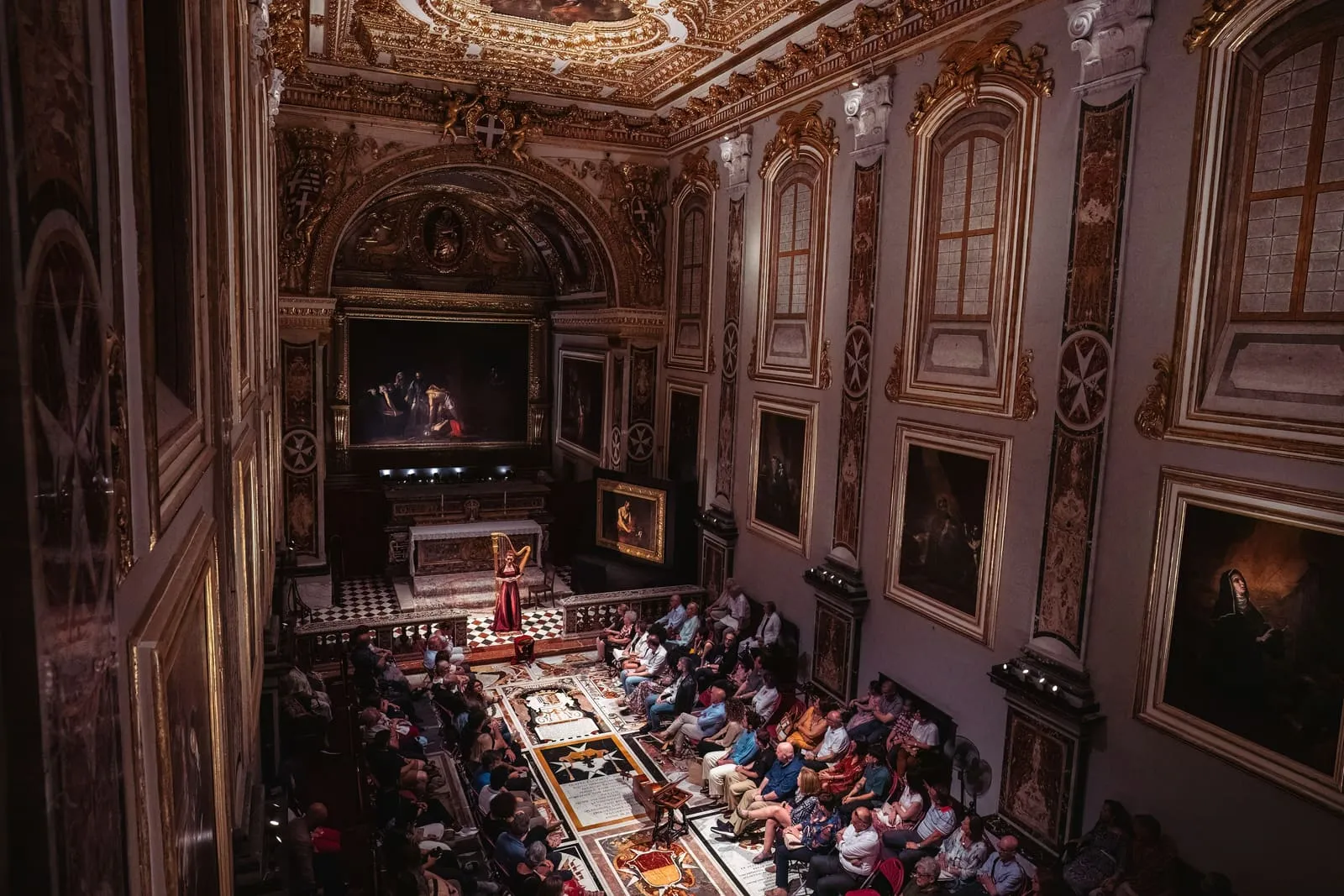 The Oratory seen from above: a harpist in a red gown performs beneath the Beheading of St John the Baptist, the audience seated around the inlaid marble floor.
