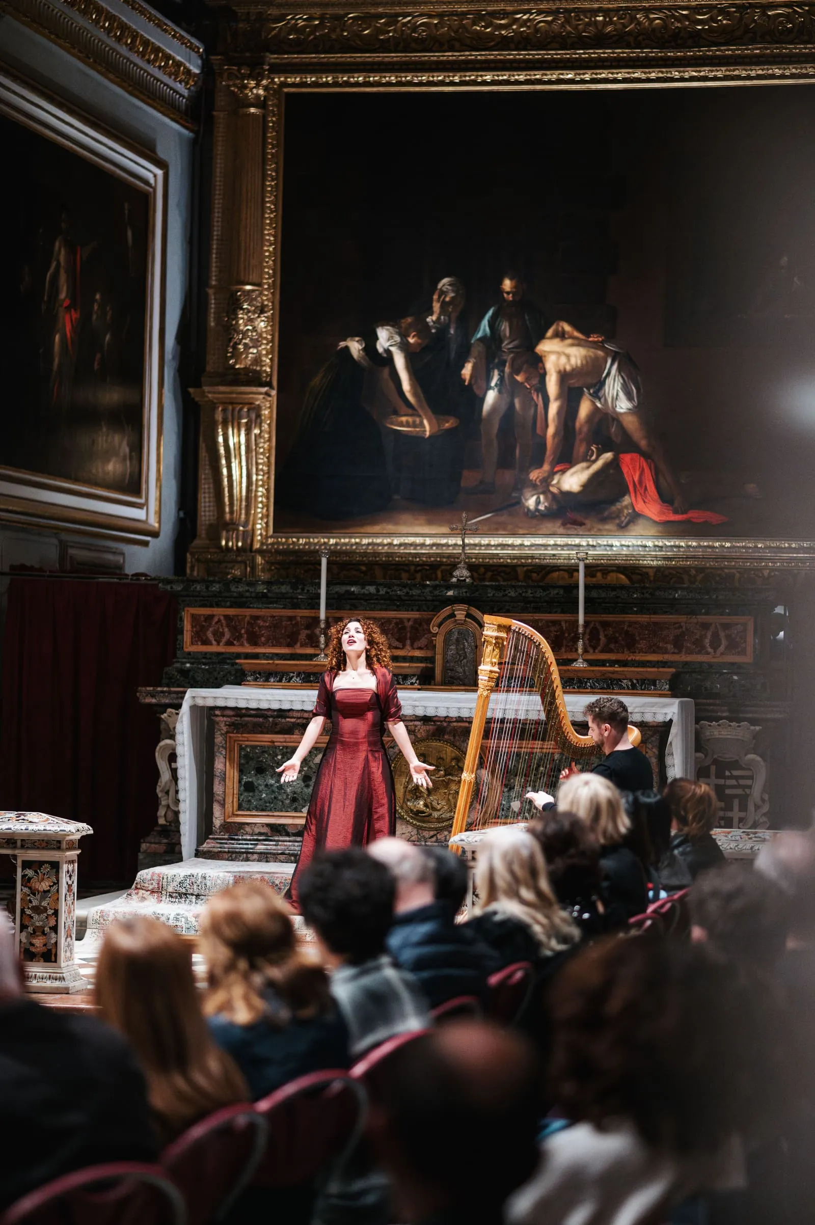 A harpist in a red gown plays a gilded Baroque harp directly beneath Caravaggio's masterpiece.
