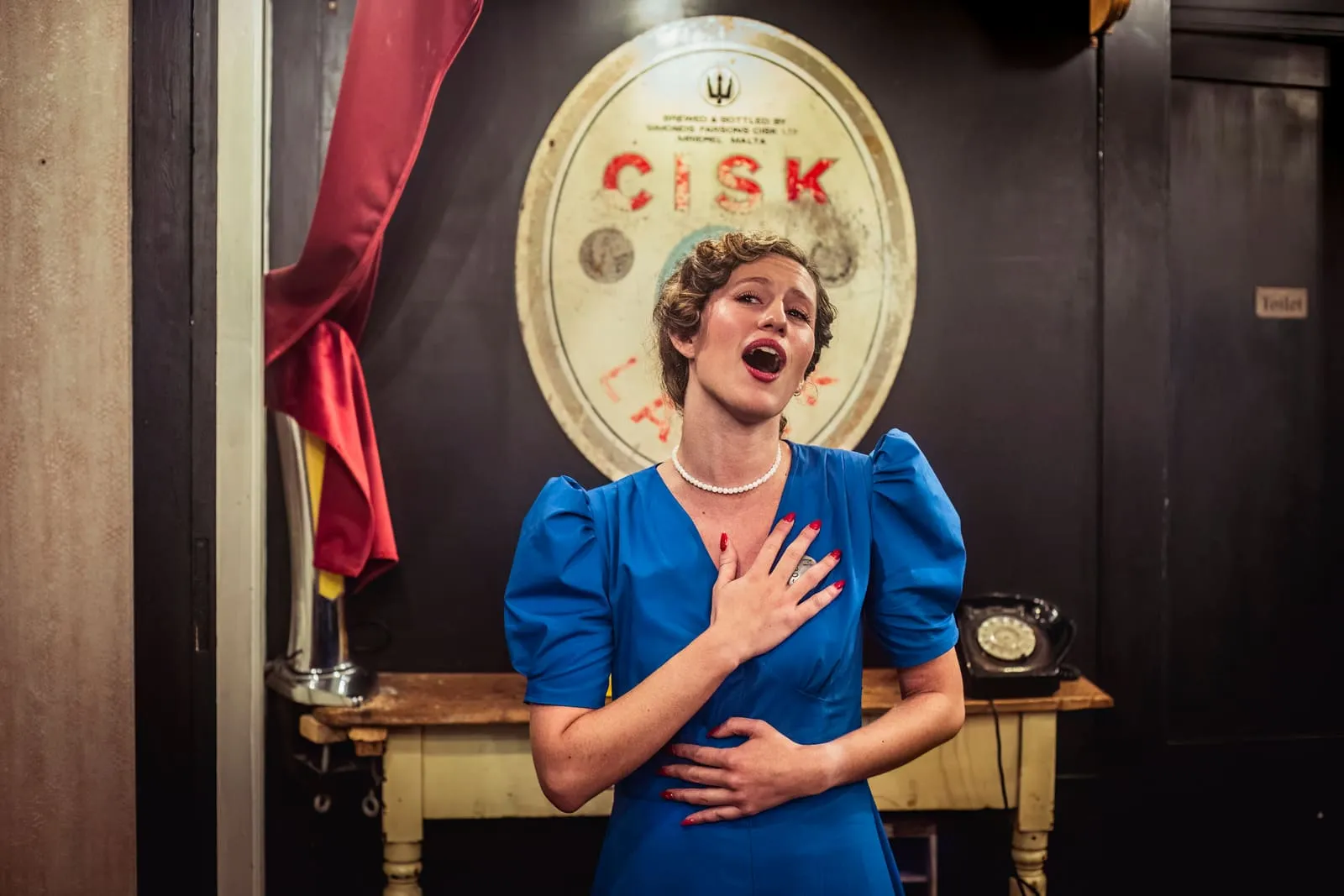 Vicietta, the singer and barmaid, singing with a hand at her heart beside the bar.