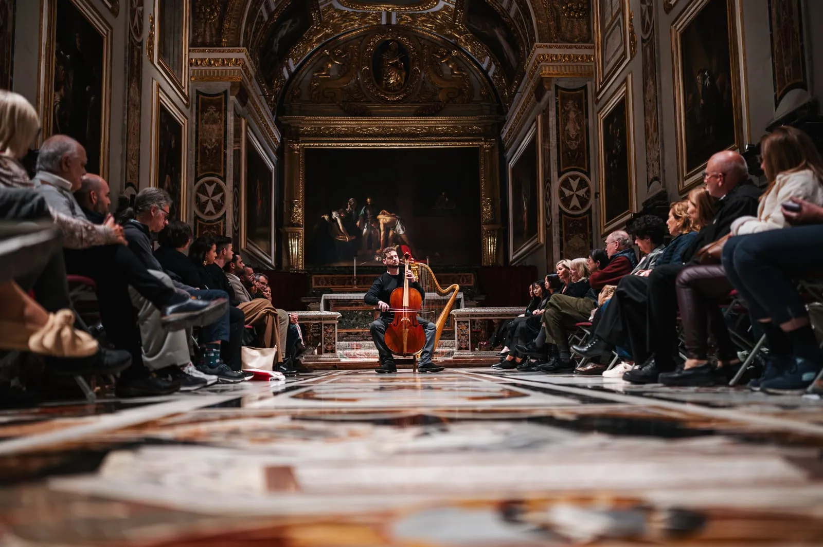 A cellist performing at the centre of the Oratory, audience seated along both sides.